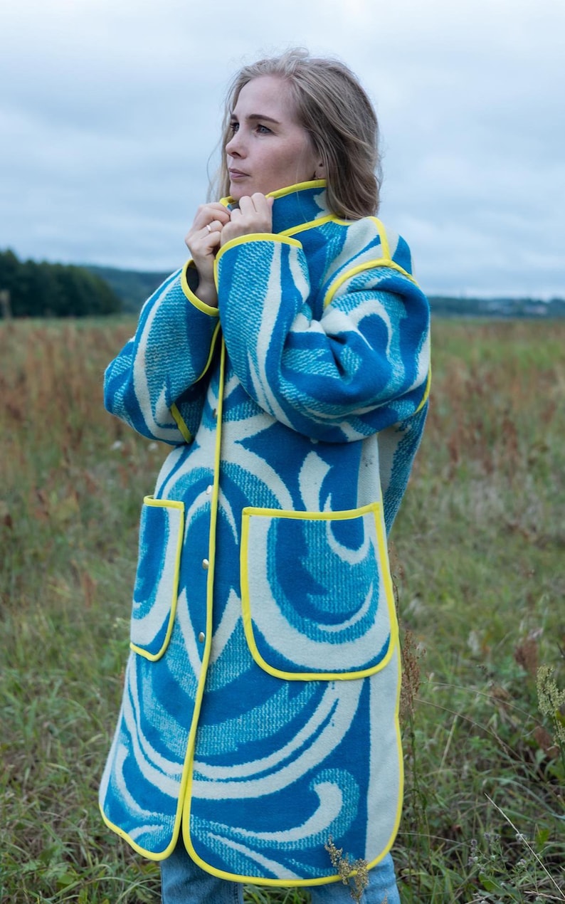 May include: A long coat with a blue and white swirl pattern and yellow trim. The coat has two front pockets and a button-up closure. The person is wearing the coat in a field.