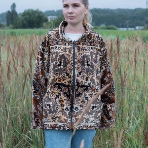 May include: A brown, black, and tan patterned zip-up jacket with a high collar. The jacket features a detailed floral design and is worn with blue jeans. The background shows a field and overcast sky.