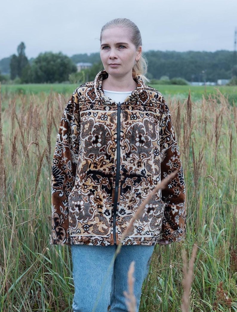 May include: A brown, black, and tan patterned zip-up jacket with a high collar. The jacket features a detailed floral design and is worn with blue jeans. The background shows a field and overcast sky.