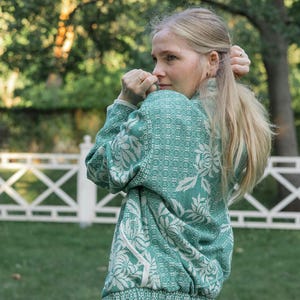 May include: A long-sleeved green and white patterned sweater featuring a floral design and a high collar, paired with light blue denim jeans. The sweater has a textured knit construction with white trim along the pockets and hemline.