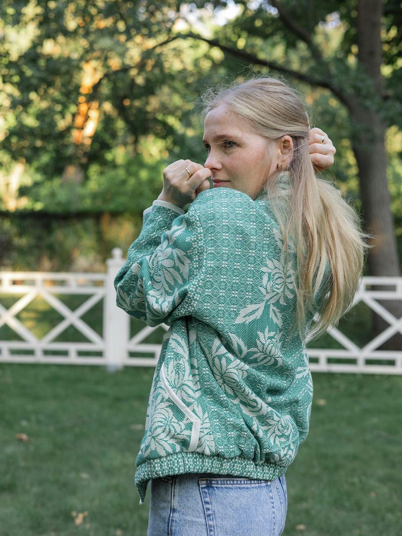 May include: A long-sleeved green and white patterned sweater featuring a floral design and a high collar, paired with light blue denim jeans. The sweater has a textured knit construction with white trim along the pockets and hemline.