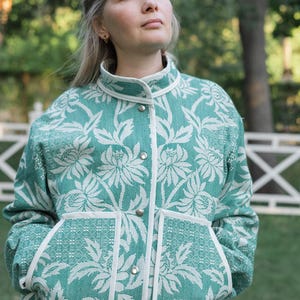 May include: A teal and white floral patterned jacket featuring a high collar, long sleeves, and a front button closure with silver-toned metal snaps. The garment includes two large front pockets with white trim and a structured, woven fabric texture.