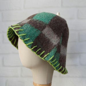 Puede incluir: Un sombrero cloche a cuadros en tonos marr&oacute;n, verde y gris, exhibido en una cabeza de maniqu&iacute;. El sombrero presenta un ribete verde ne&oacute;n cosido alrededor del borde. El dise&ntilde;o del sombrero es un patr&oacute;n de cuadros cl&aacute;sico.
