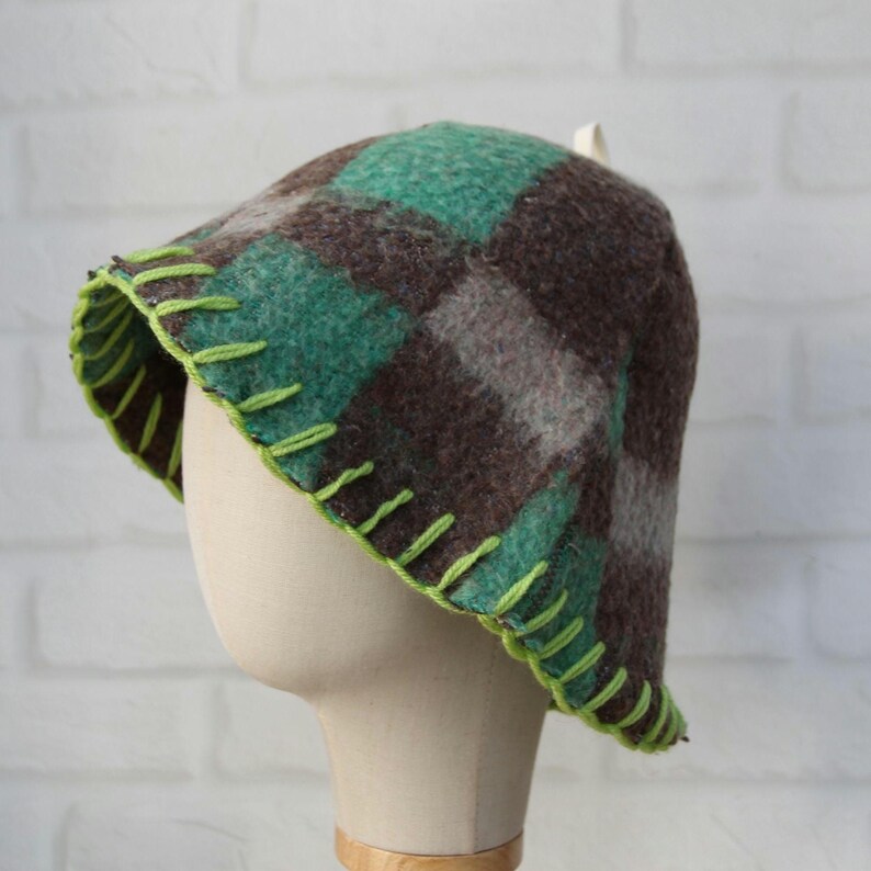 Puede incluir: Un sombrero cloche a cuadros en tonos marr&oacute;n, verde y gris, exhibido en una cabeza de maniqu&iacute;. El sombrero presenta un ribete verde ne&oacute;n cosido alrededor del borde. El dise&ntilde;o del sombrero es un patr&oacute;n de cuadros cl&aacute;sico.