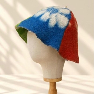 Może przedstawiać: Filcowy kapelusz typu bucket hat o żywym wzorze typu color-block. Kapelusz posiada duży wzór białego kwiatu na niebieskim tle, z jednolitym zielonym panelem po jednej stronie i jednolitym czerwonym panelem po drugiej stronie ronda.