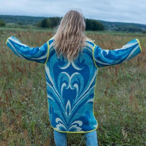 May include: A long, open-front cardigan in shades of blue and white with a decorative pattern. The cardigan has yellow trim and is worn over blue jeans. The person is standing in a field with arms outstretched.