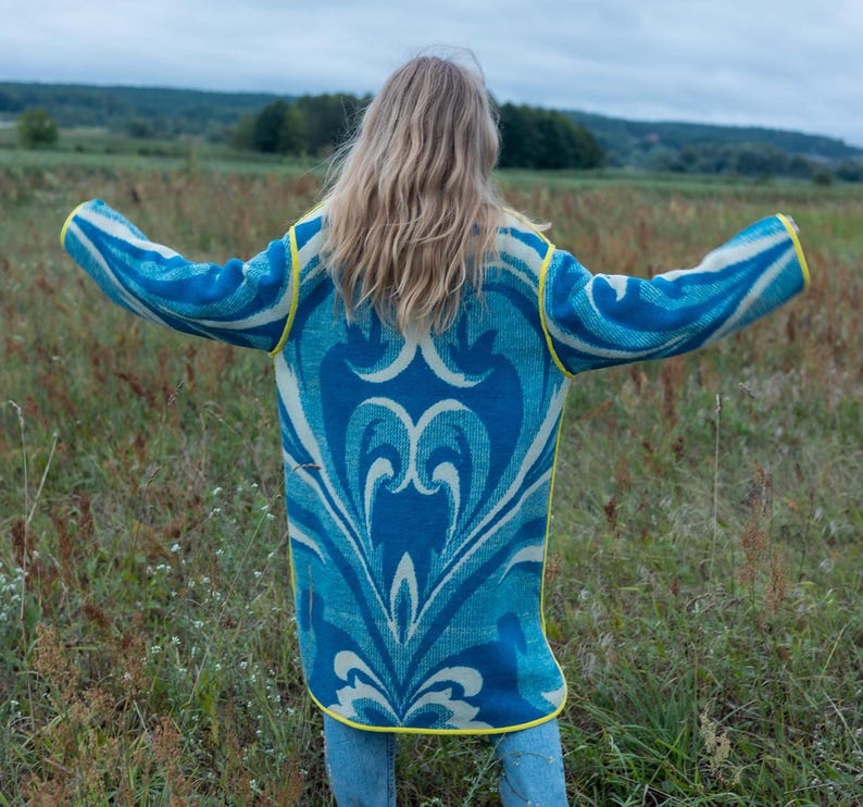 May include: A long, open-front cardigan in shades of blue and white with a decorative pattern. The cardigan has yellow trim and is worn over blue jeans. The person is standing in a field with arms outstretched.
