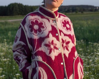 May include: A burgundy jacket with a floral pattern in cream. The jacket has a stand-up collar, button closure, and cream-colored trim. The jacket is worn over a light-colored top and blue jeans. The background is a field of wildflowers.
