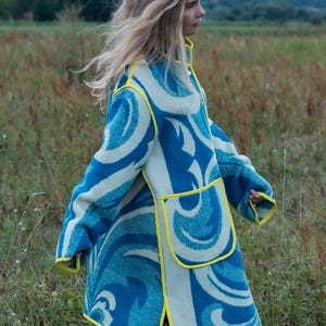 May include: A long coat with a blue and white wave pattern and yellow trim. The coat has two large pockets and is worn over blue jeans. The coat is made of a thick material, suitable for cooler weather.