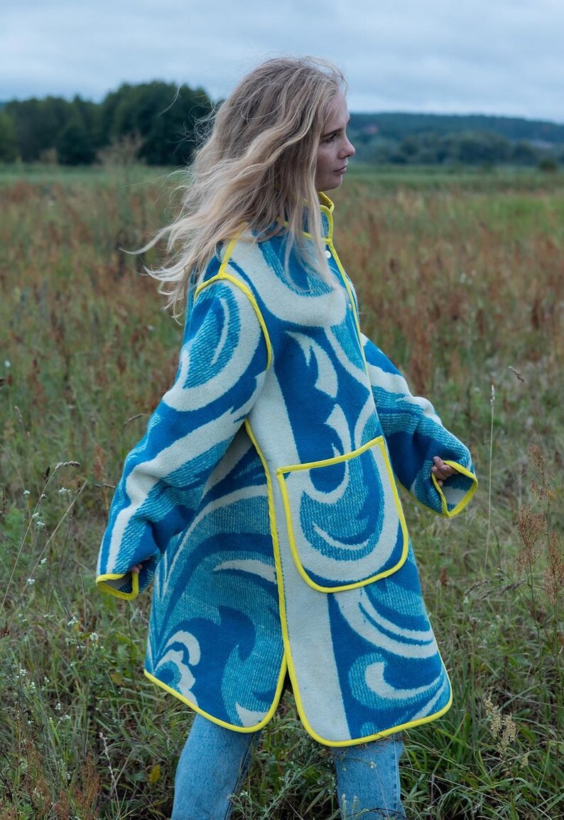 May include: A long coat with a blue and white wave pattern and yellow trim. The coat has two large pockets and is worn over blue jeans. The coat is made of a thick material, suitable for cooler weather.