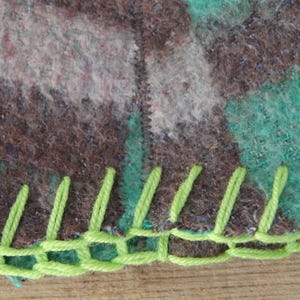 Puede incluir: Primer plano de una secci&oacute;n de una manta con un patr&oacute;n de camuflaje en tonos marr&oacute;n, gris y verde azulado. El borde est&aacute; acabado con costuras de color verde brillante, creando un borde decorativo. La manta parece estar hecha de un material similar a la lana.