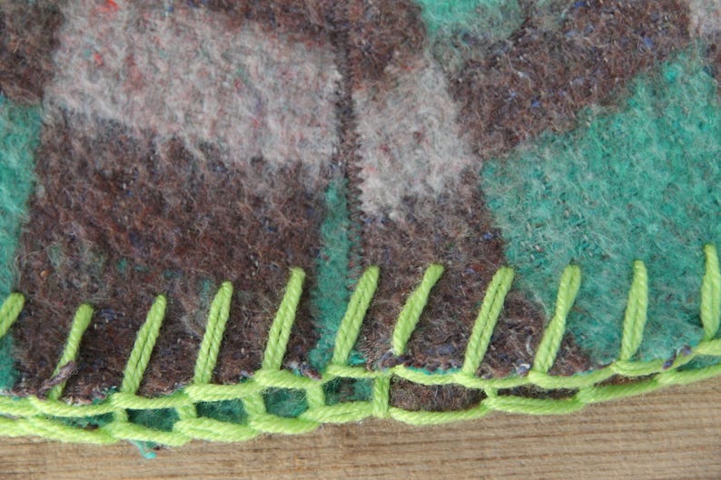 Puede incluir: Primer plano de una secci&oacute;n de una manta con un patr&oacute;n de camuflaje en tonos marr&oacute;n, gris y verde azulado. El borde est&aacute; acabado con costuras de color verde brillante, creando un borde decorativo. La manta parece estar hecha de un material similar a la lana.