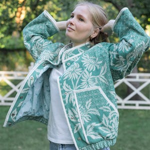 May include: A teal and white floral patterned jacket with long sleeves, a stand-up collar, and white trim along the edges. The jacket features a front snap-button closure, two front pockets, and a light blue interior lining, worn over a white shirt.
