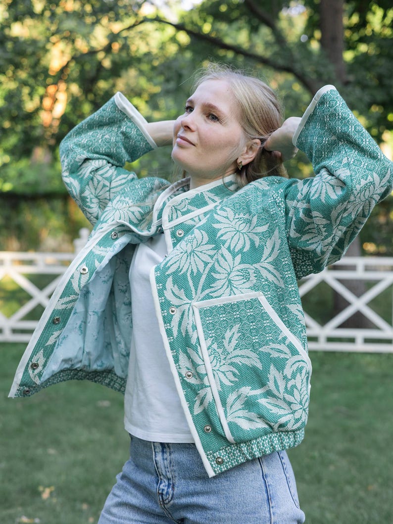 May include: A teal and white floral patterned jacket with long sleeves, a stand-up collar, and white trim along the edges. The jacket features a front snap-button closure, two front pockets, and a light blue interior lining, worn over a white shirt.