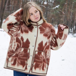 Puede incluir: Chaqueta beige y marrón estampada con cuello alto y dos bolsillos delanteros. La chaqueta tiene cierre de botones y ribete marrón. La modelo lleva vaqueros azules. El fondo es un bosque nevado.