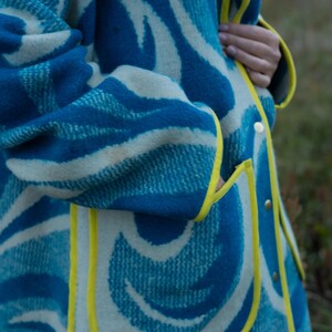 May include: A blue and white patterned coat with yellow trim and gold buttons. The coat features a unique wave-like design in shades of blue and white. The coat has a pocket and is made of a thick, textured material.