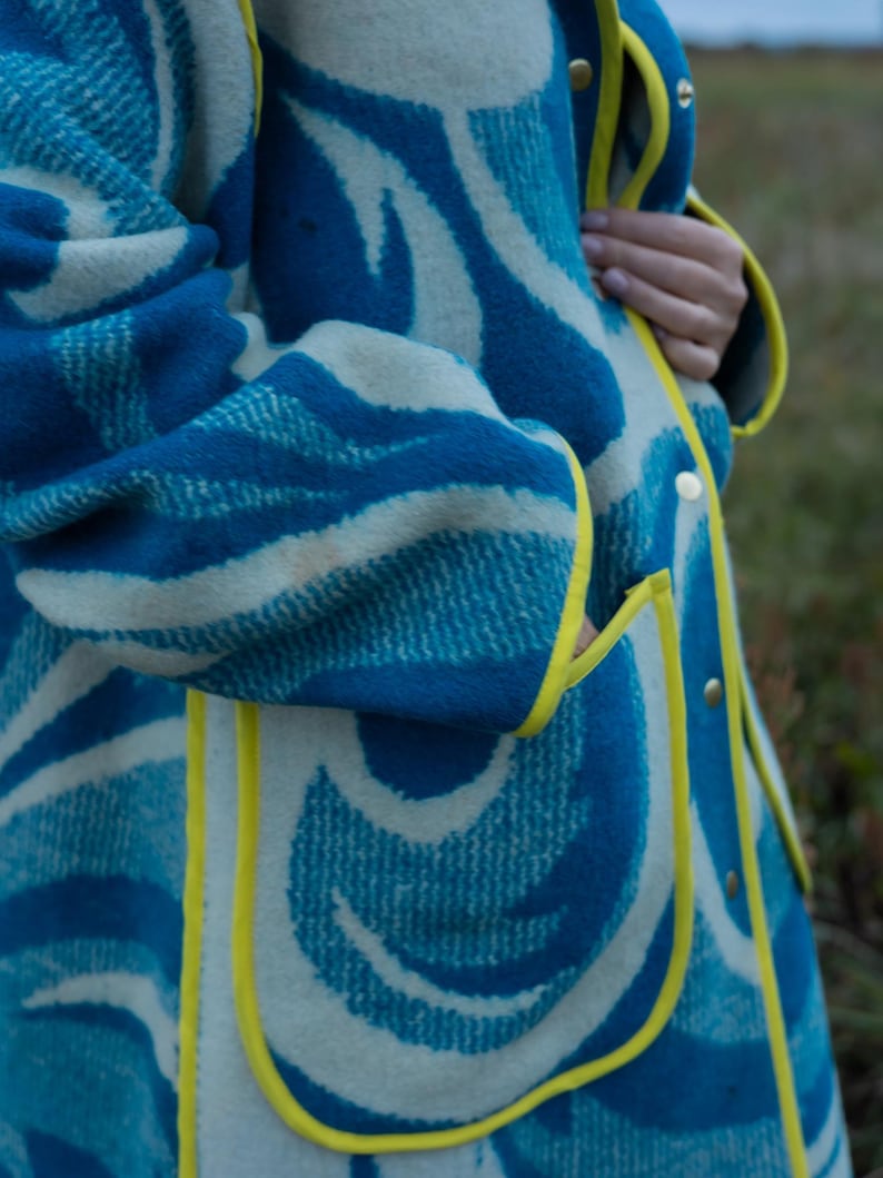 May include: A blue and white patterned coat with yellow trim and gold buttons. The coat features a unique wave-like design in shades of blue and white. The coat has a pocket and is made of a thick, textured material.