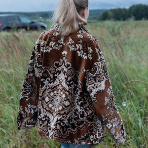 May include: A brown patterned jacket with a floral design is worn by a person walking through a field of tall grass. The jacket has a button-up front and long sleeves. The person is wearing blue jeans.