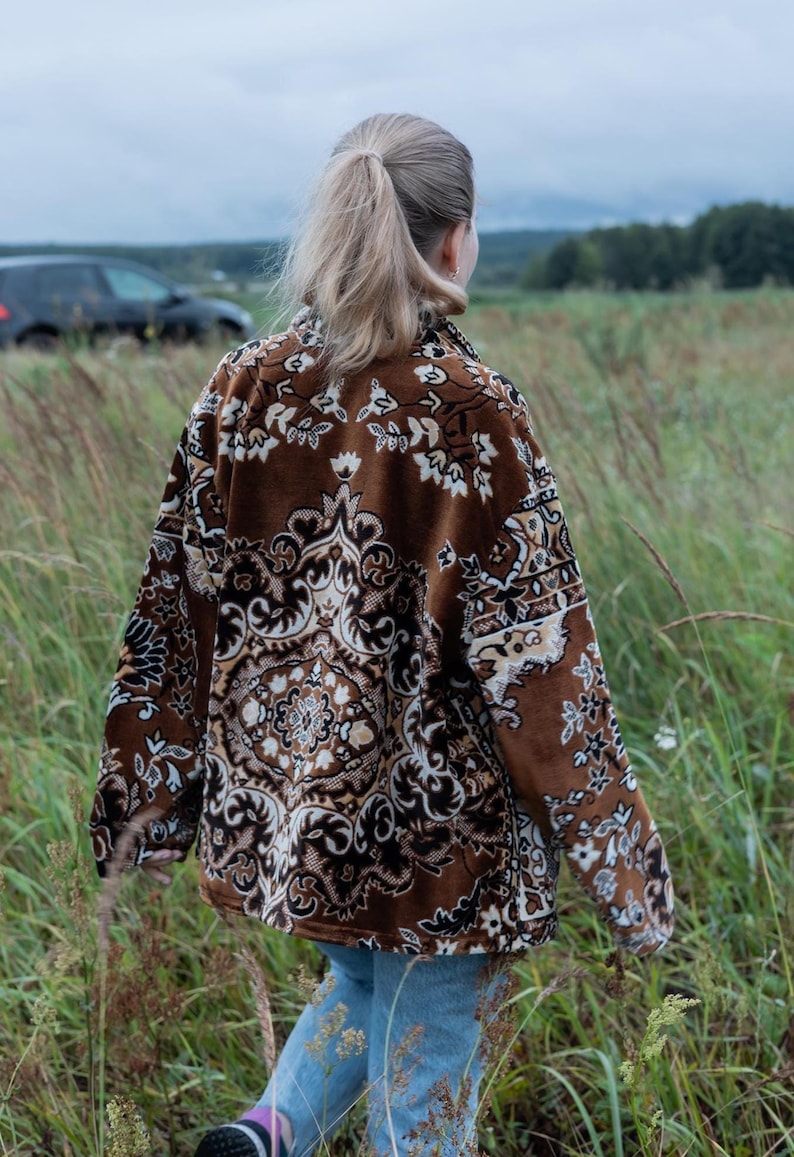 May include: A brown patterned jacket with a floral design is worn by a person walking through a field of tall grass. The jacket has a button-up front and long sleeves. The person is wearing blue jeans.