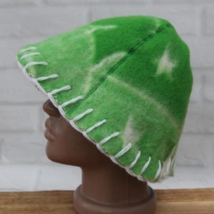 Peut inclure: Un bonnet de sauna en feutre vert avec des coutures blanches sur le bord. Le chapeau présente un motif tie-dye vert clair et blanc. Le chapeau est présenté sur une tête de mannequin marron.