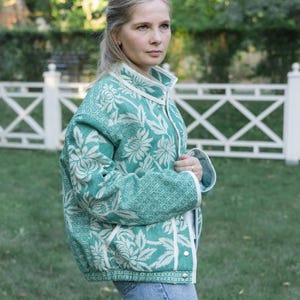 May include: A teal and white floral patterned jacket with a high collar, long sleeves, and silver-toned metal snap buttons down the front. The garment features a textured woven fabric with a repeating botanical design and white trim along the edges.