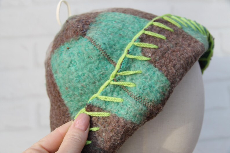 Puede incluir: Un sombrero hecho a mano con un patr&oacute;n de cuadros en tonos marrones y verde azulado. El sombrero presenta costuras de color verde brillante a lo largo del borde. El sombrero est&aacute; hecho de un material suave y texturizado.