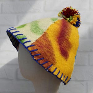 May include: A colorful, patchwork hat with a pom-pom. The hat features a cream-colored section with green polka dots, and a yellow, orange, and red striped section. The brim is stitched with blue thread. A multi-colored pom-pom tops the hat.