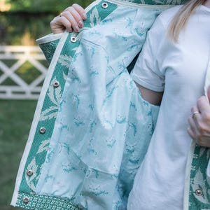 May include: A jacket featuring a green and white woven leaf pattern on the exterior with a light blue interior lining decorated with a small green botanical print. The garment includes a white trim along the edges and silver metal snap buttons.