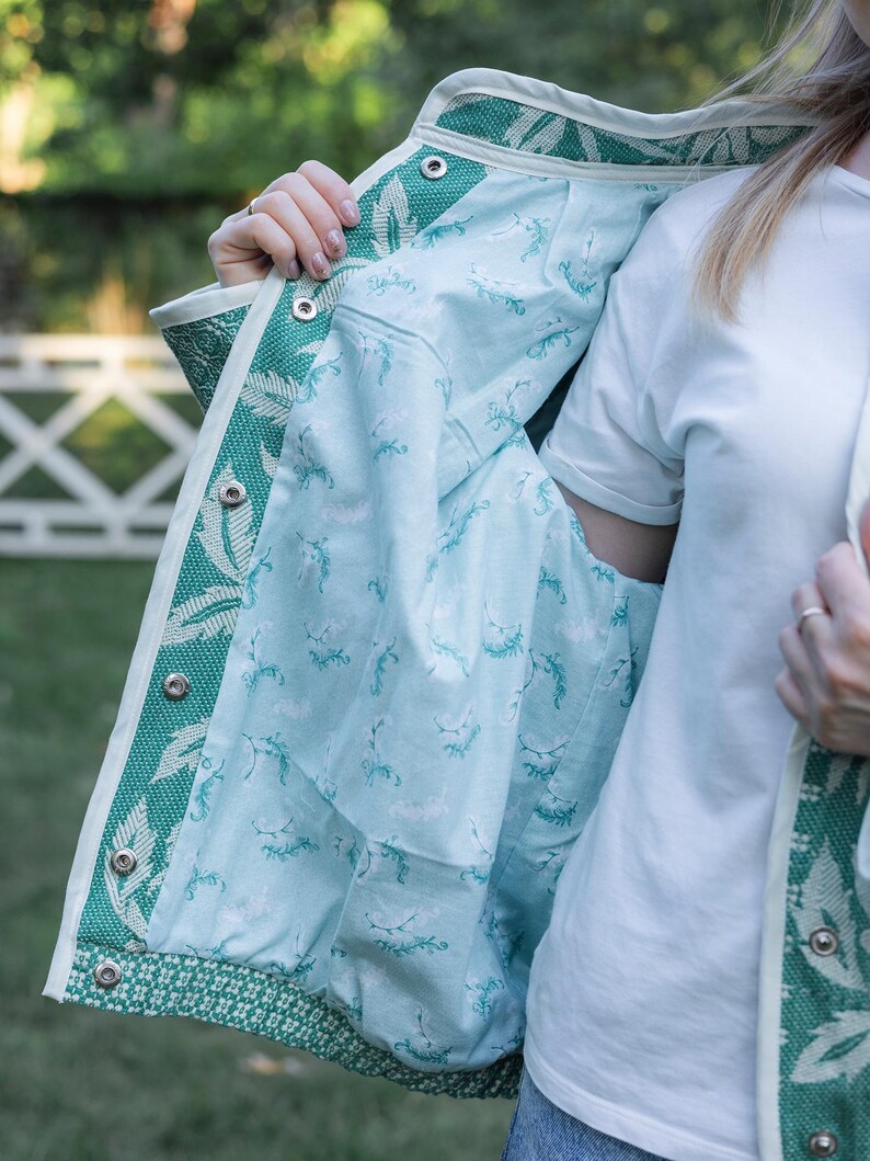 May include: A jacket featuring a green and white woven leaf pattern on the exterior with a light blue interior lining decorated with a small green botanical print. The garment includes a white trim along the edges and silver metal snap buttons.
