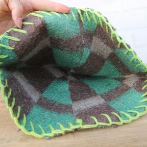 Puede incluir: Una bolsa hecha a mano, abierta por arriba, con un estampado de cuadros en tonos verdes, marrones y grises. Los bordes est&aacute;n cosidos con hilo verde brillante. La bolsa est&aacute; hecha de un material suave, posiblemente afieltrado.