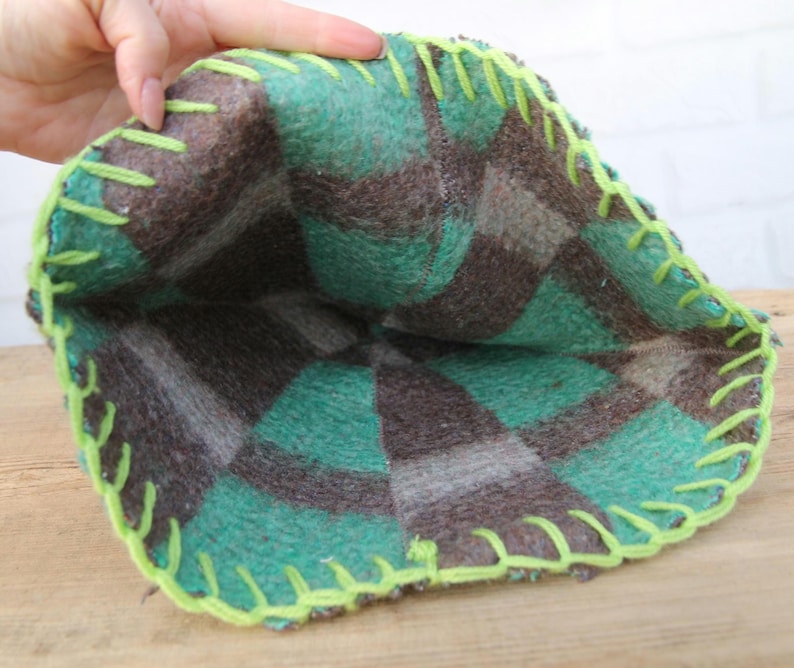 Puede incluir: Una bolsa hecha a mano, abierta por arriba, con un estampado de cuadros en tonos verdes, marrones y grises. Los bordes est&aacute;n cosidos con hilo verde brillante. La bolsa est&aacute; hecha de un material suave, posiblemente afieltrado.