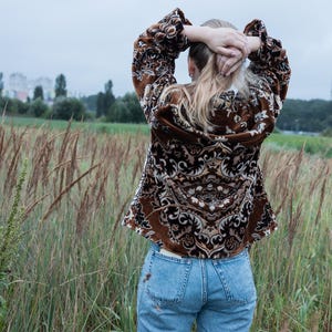 May include: A brown patterned jacket with a detailed design, paired with light blue jeans. The jacket features a repeating pattern in shades of brown and cream. The person is standing in a field of tall grass, with their hands behind their head.