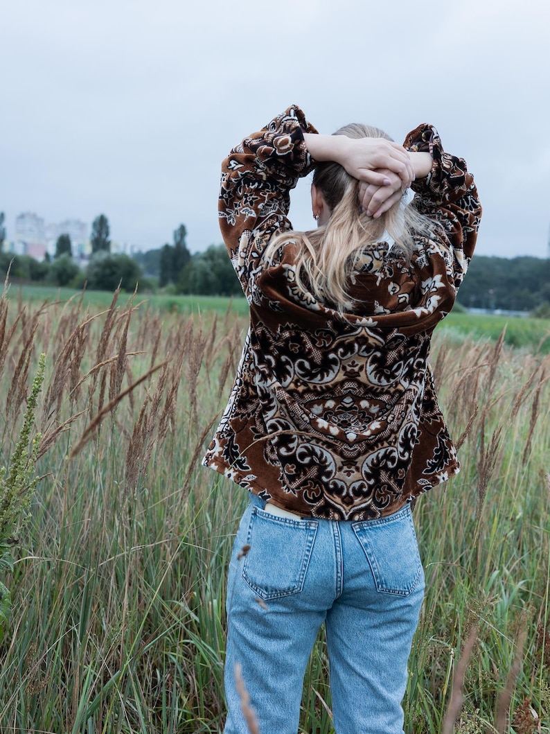 May include: A brown patterned jacket with a detailed design, paired with light blue jeans. The jacket features a repeating pattern in shades of brown and cream. The person is standing in a field of tall grass, with their hands behind their head.