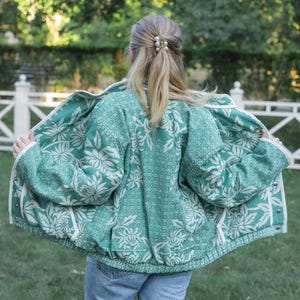 May include: A teal and white floral patterned jacket with a textured weave, long sleeves, and a front button closure. The jacket features a repeating botanical print across the back and interior panels, with white trim along the edges and cuffs.