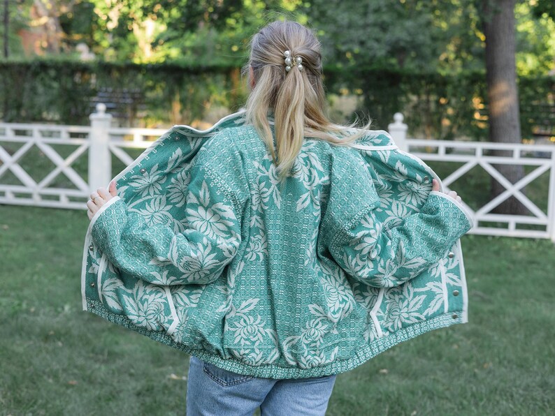 May include: A teal and white floral patterned jacket with a textured weave, long sleeves, and a front button closure. The jacket features a repeating botanical print across the back and interior panels, with white trim along the edges and cuffs.
