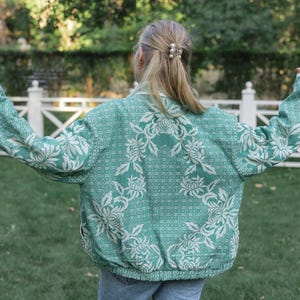 May include: A long-sleeved jacket in a vibrant teal green color featuring a prominent white floral pattern throughout the fabric. The jacket has a structured collar, a full-length front zipper, and a ribbed hemline at the waist and cuffs.