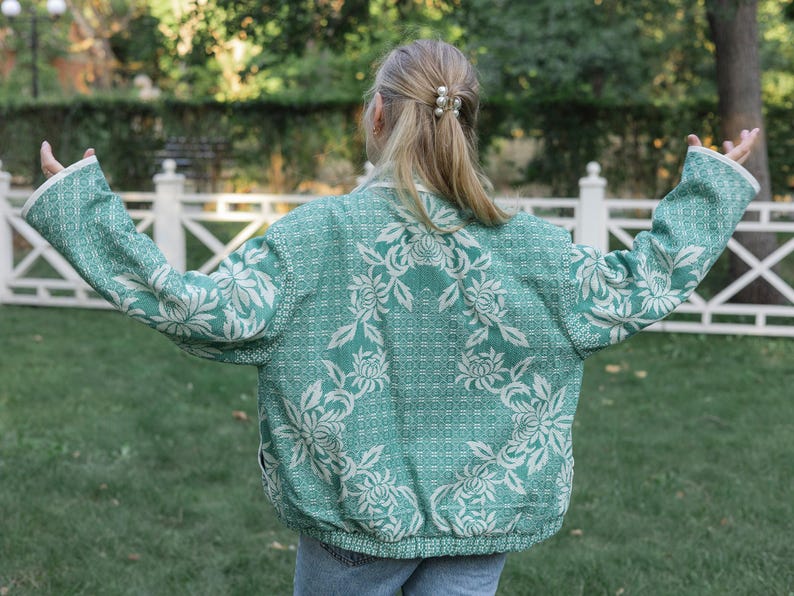 May include: A long-sleeved jacket in a vibrant teal green color featuring a prominent white floral pattern throughout the fabric. The jacket has a structured collar, a full-length front zipper, and a ribbed hemline at the waist and cuffs.