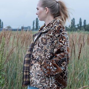 May include: A brown and beige patterned jacket with a floral design. The jacket has a zipper closure and a plaid lining. The person is wearing blue jeans and is standing in a field of tall grass.