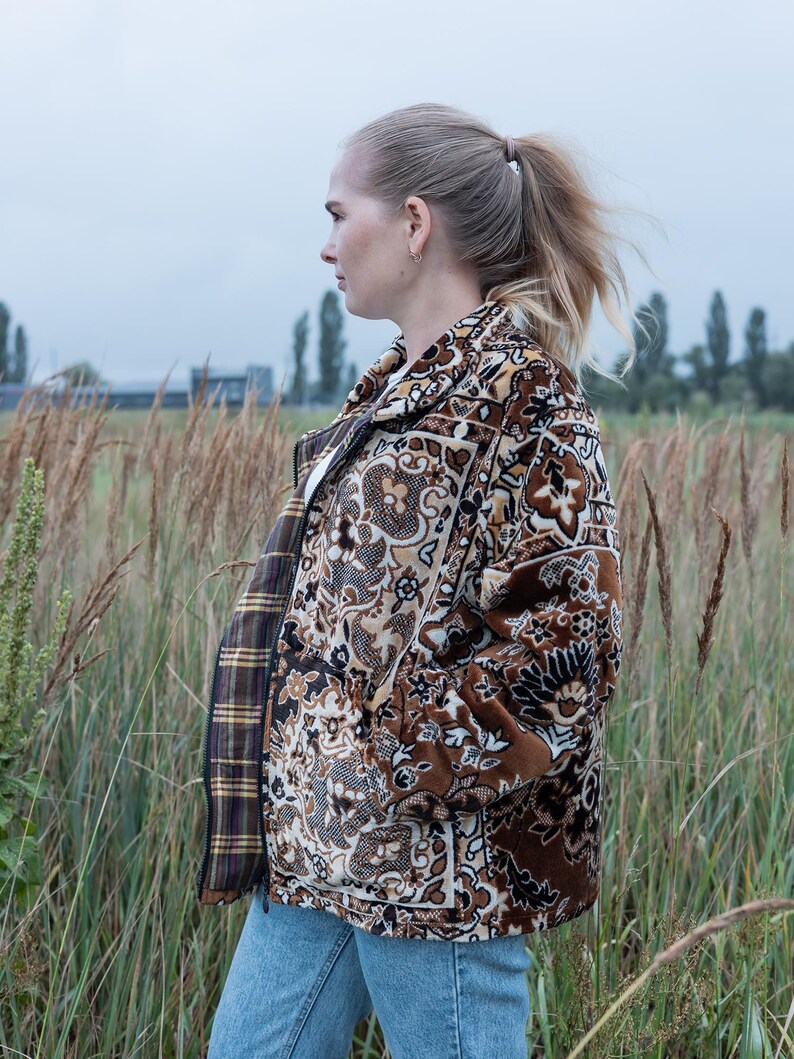 May include: A brown and beige patterned jacket with a floral design. The jacket has a zipper closure and a plaid lining. The person is wearing blue jeans and is standing in a field of tall grass.