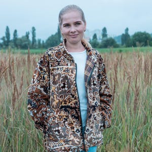 May include: A brown patterned jacket with a zipper, worn over a white shirt and blue jeans. The jacket features a detailed floral and geometric design in shades of brown, black, and beige. The person is standing in a field of tall grass.