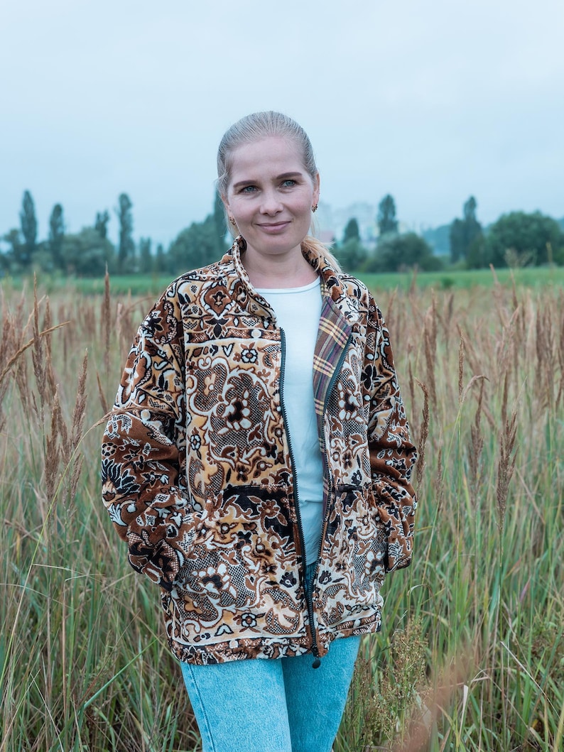 May include: A brown patterned jacket with a zipper, worn over a white shirt and blue jeans. The jacket features a detailed floral and geometric design in shades of brown, black, and beige. The person is standing in a field of tall grass.