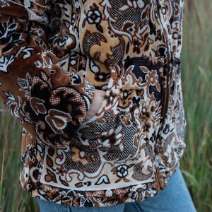 May include: A brown and beige patterned jacket with a zipper closure and pockets. The jacket features a floral and geometric design. The person is wearing blue jeans. The jacket is made of a soft, textured material.