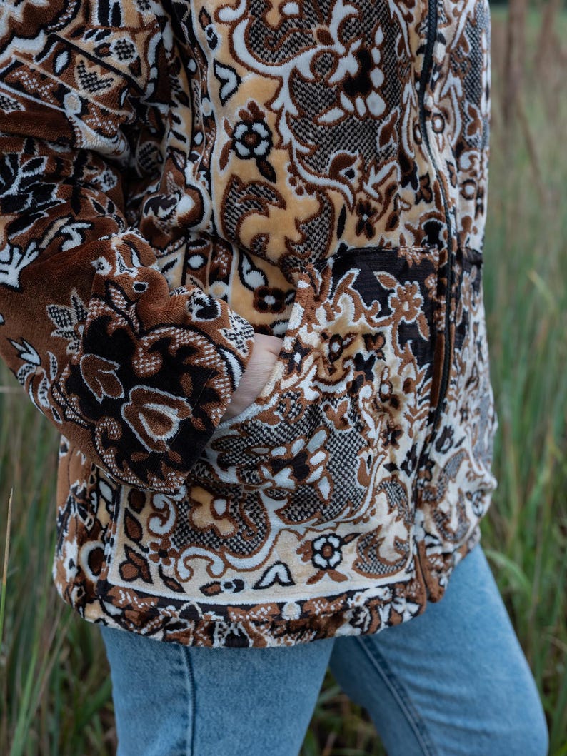 May include: A brown and beige patterned jacket with a zipper closure and pockets. The jacket features a floral and geometric design. The person is wearing blue jeans. The jacket is made of a soft, textured material.