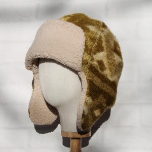 May include: A winter hat with ear flaps, displayed on a mannequin head. The hat features a patterned olive green and tan exterior with a soft, cream-colored lining and ear flaps. The hat is designed to provide warmth and protection from the cold.