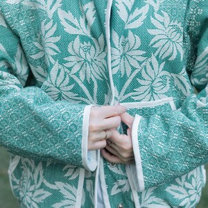May include: A teal and white jacket featuring a repeating floral pattern with large stylized blossoms and leaves. The garment has long sleeves, a front opening with a white trim, and two front pockets, all finished with a clean white edge binding.