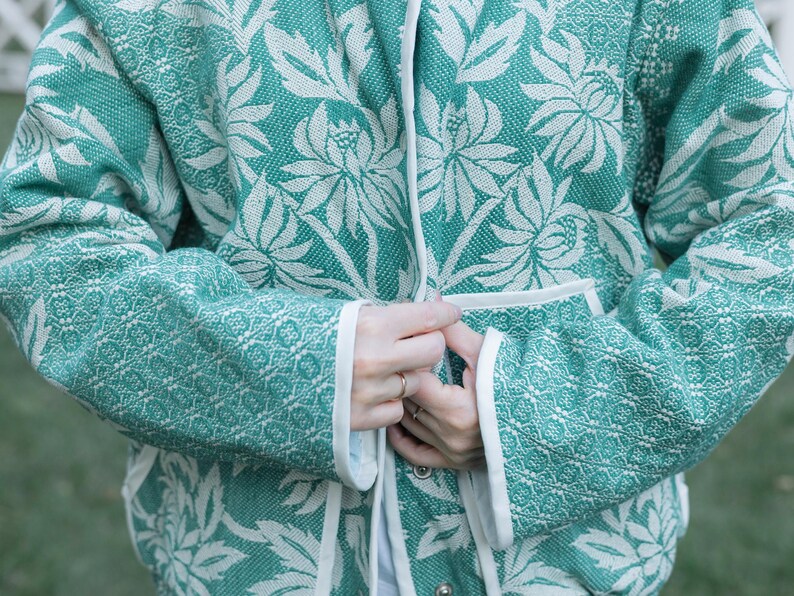 May include: A teal and white jacket featuring a repeating floral pattern with large stylized blossoms and leaves. The garment has long sleeves, a front opening with a white trim, and two front pockets, all finished with a clean white edge binding.