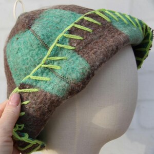 Puede incluir: Un sombrero hecho a mano con un patr&oacute;n de cuadros en tonos marrones y verde azulado. El borde est&aacute; acentuado con costuras verde brillante. El sombrero se exhibe en una cabeza de maniqu&iacute;.