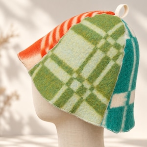 May include: A felted sauna hat featuring a patchwork design of orange, light green, and dark teal plaid patterns. The hat has a conical shape with visible white stitching along the seams and a small white fabric loop at the very top peak.