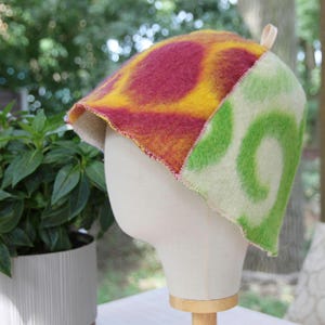 May include: A felted hat featuring a vibrant pattern of large magenta and yellow circular shapes on one side and a light cream base with bright green swirling designs on the other. The hat has a small loop at the top and a raw, stitched edge.