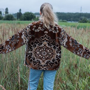 May include: A brown jacket with an ornate, repeating pattern in beige and black. The jacket is worn over blue jeans. The person is standing in a field of tall grass with a background of trees and a cloudy sky.
